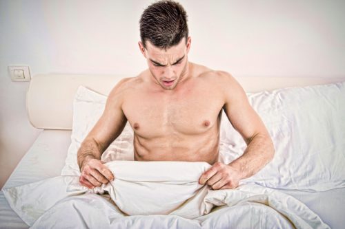 surprised half naked young man in bed looking down at his underwear at his penis under white covers sheet in badroom. Concept photo of male sexuality and man sex problems, domestic atmosphere.